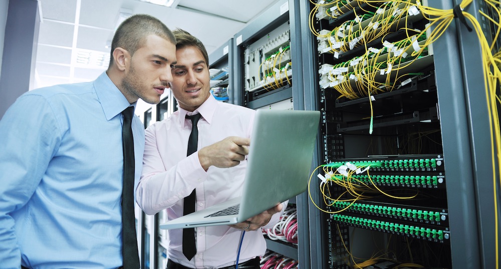 Two IT professionals in a server room looking at a laptop